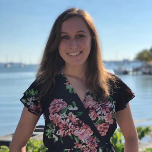 Irene, a white, Jewish woman, smiles at the camera on a sunny day by the water. She has long brown hair and wears a black  with pink flowers on it.