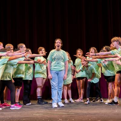 A group of kids stand in an inverted triangle, motions towards one student in the middle
