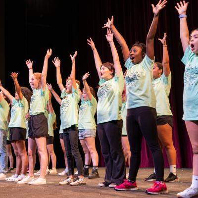 Kids in a line sing with their arms stretched towards the sky 