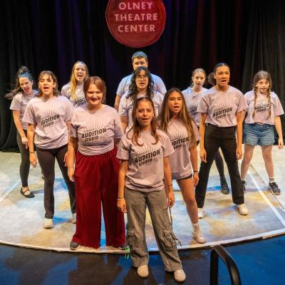 A group of high schoolers in a triangle formation, singing to the audience 