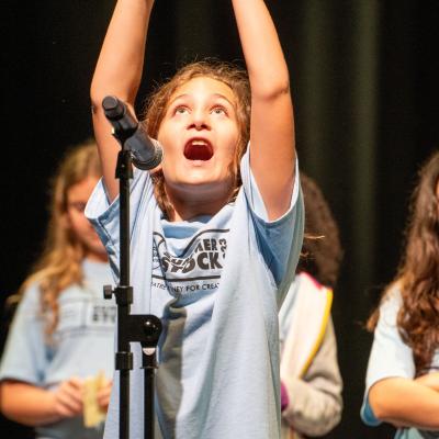 A young kid with their arms in the air, looking up, and singing into the microphone