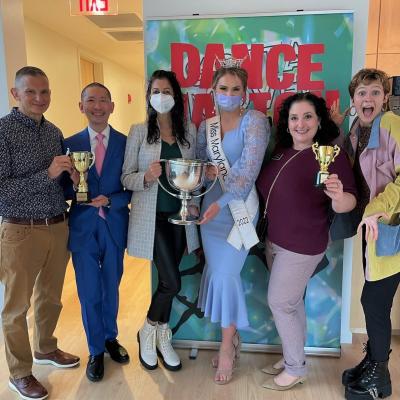 Six people pose with trophies in front of a green banner with red lettering that reads "Dance Nation". In the center are Dr. Chandni Shah and "Miss Maryland" Kayla Willing.