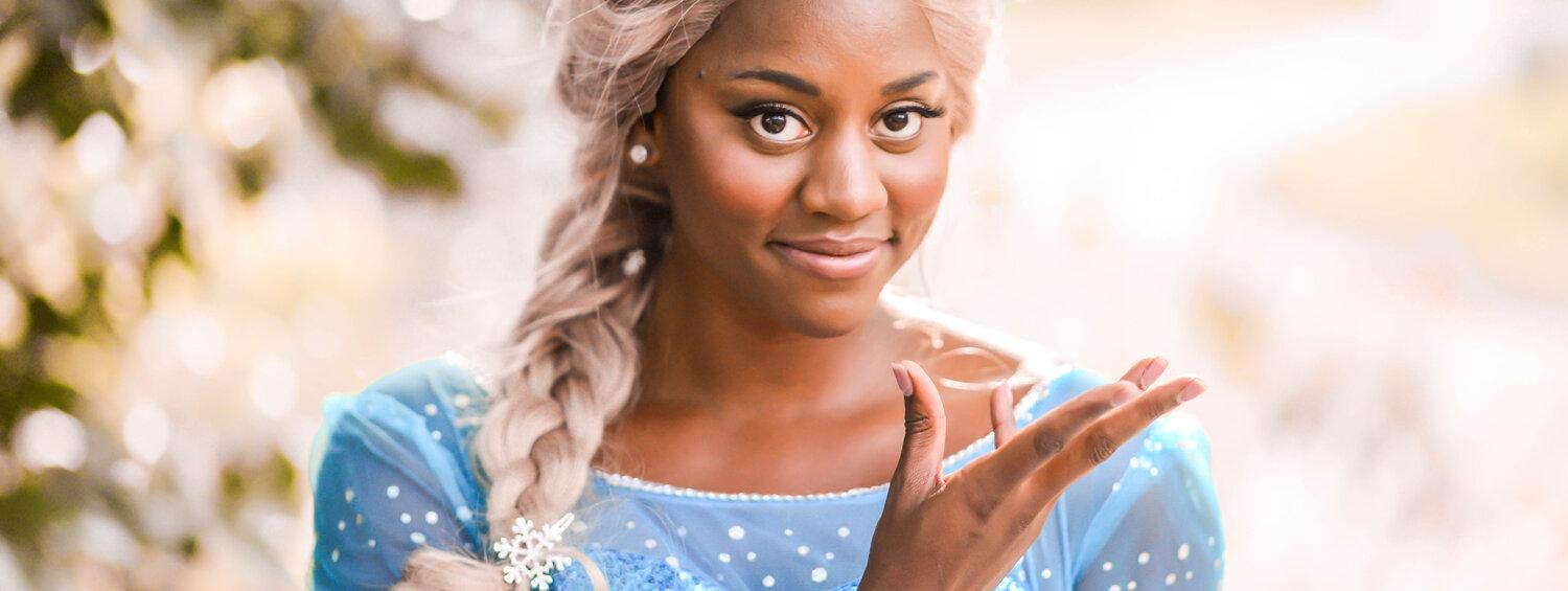 Simone Brown is dressed as the "Ice Queen" (aka. Princess Elsa) in a light blue sparkly gown and blonde wig. She has  warm brown skin, deep brown eyes, rosy cheeks, and smiles softly at the camera.