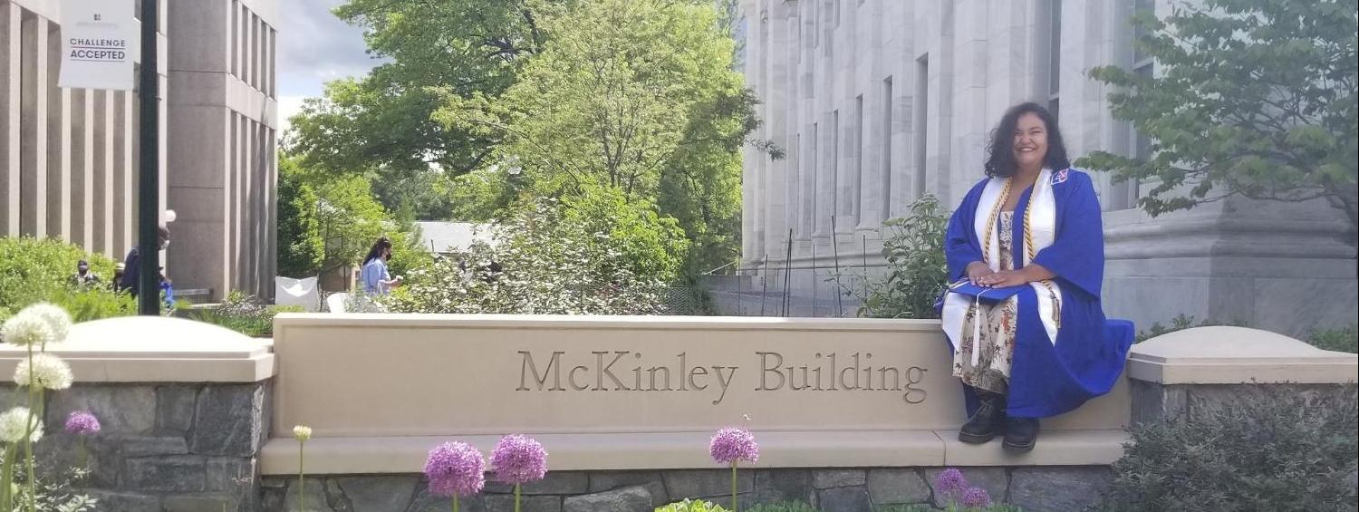 Image of Sultana Qureshi in blue graduation robes sitting on college sign "Mckinley Building"