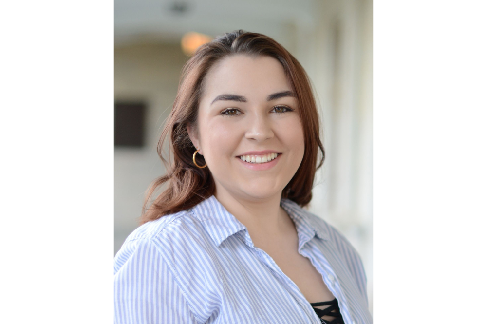 Cat, a young white woman with long brown hair and small hoop earrings, smiles at the camera. She wears a light blue button-down shirt.