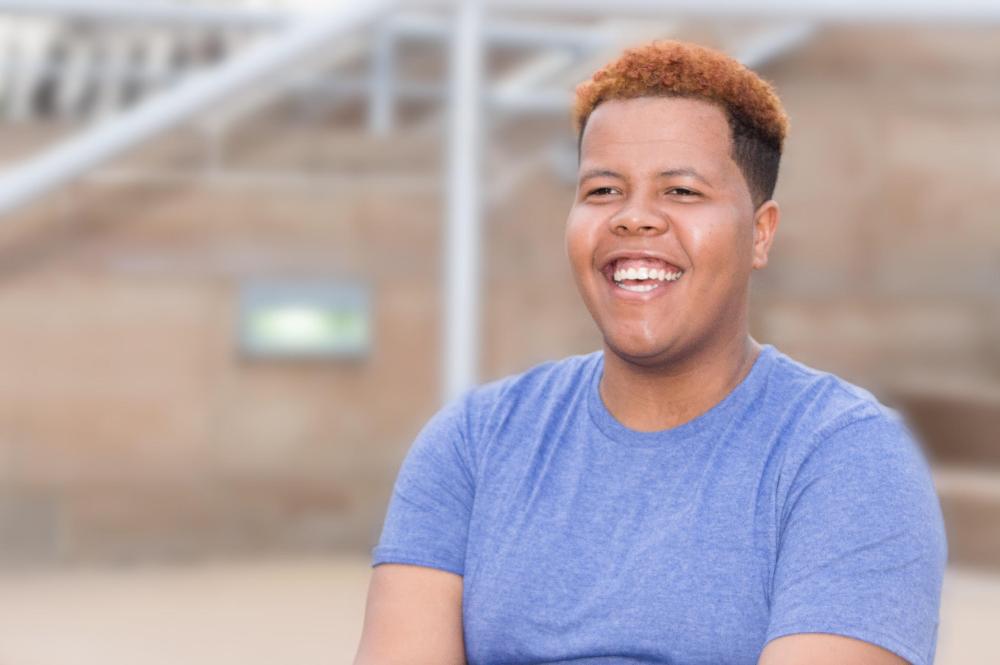 Rashaud smiles just past the camera's eye. They have medium brown skin, short, kinky curls died ombre on top, and a sky blue shirt. A blurred background of outdoor architecture is behind him.