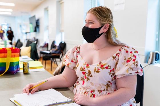 Image of Dramaturg Alissa Klusky (masked) sitting at a table with the script for The Joy that Carries You, she is smiling