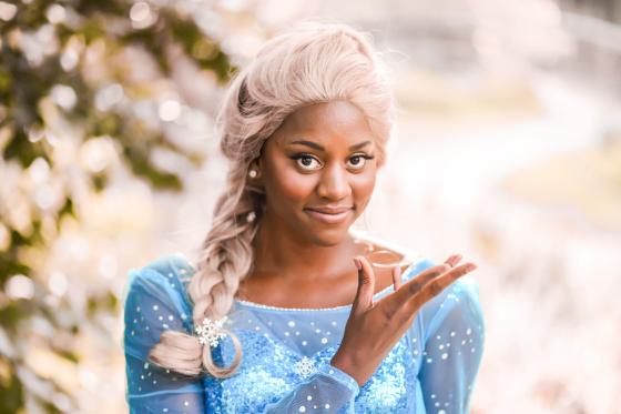 Simone Brown is dressed as the "Ice Queen" (aka. Princess Elsa) in a light blue sparkly gown and blonde wig. She has  warm brown skin, deep brown eyes, rosy cheeks, and smiles softly at the camera.