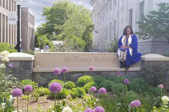 Image of Sultana Qureshi in blue graduation robes sitting on college sign "Mckinley Building"