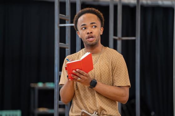 Long Way Down in rehearsal | Man in a tan shirt holding a red book 